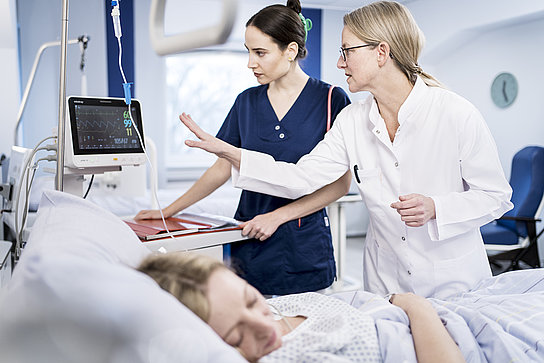 Eine Krankenschwester und eine Ärztin prüfen gemeinsam die Werte eines medizinischen Geräts einer Patientin, die im Bett liegt.