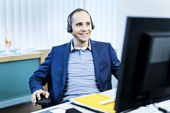 Ein Mann trägt ein Headset und blickt auf seinen Monitor. Er ist im Gespräch mit jemanden.