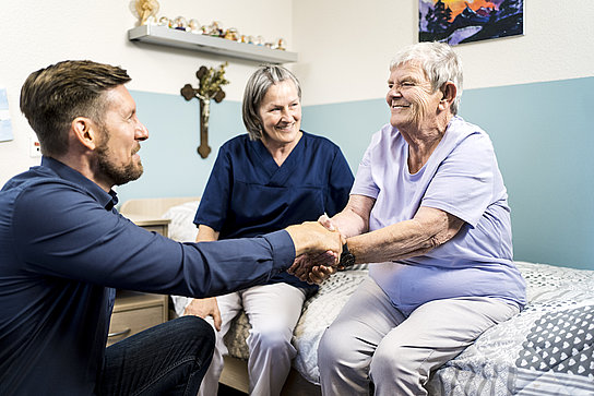 Ein Gutachter schüttelt einer Frau in ihrem Zimmer in einer Pflegeeinrichtung die Hand. Neben der Frau auf dem Bett sitzt eine Pflegerin. 