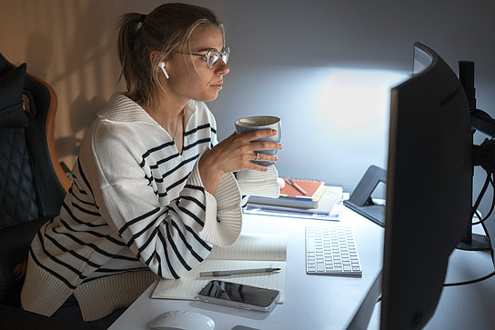 Eine Frau sitzt vor einem Monitor an ihrem Schreibtisch und trägt Kopfhörer. Sie hält eine Tasse in der Hand. Vor ihr liegen Unterlagen und ein Stift. Sie befindet sich im Home Office.