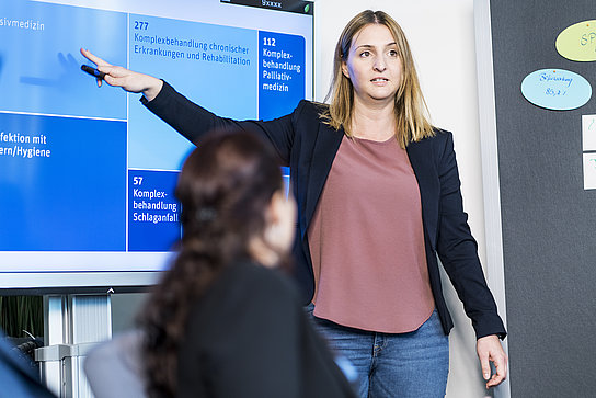 Eine Frau steht vor einem Bildschirm, auf dem eine Präsentation zu sehen ist. Sie präsentiert, was auf dem Monitor zu sehen ist. Vor ihr sitzt eine Person und schaut ihr dabei zu.